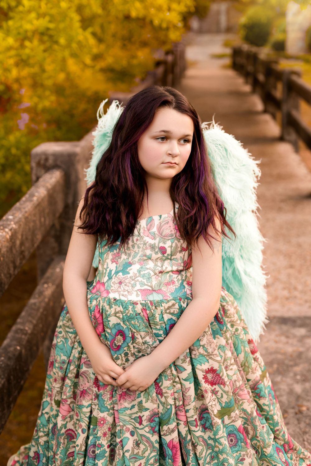 Young girl in floral dress with mint angel wings poses on a wooden bridge surrounded by autumn foliage.