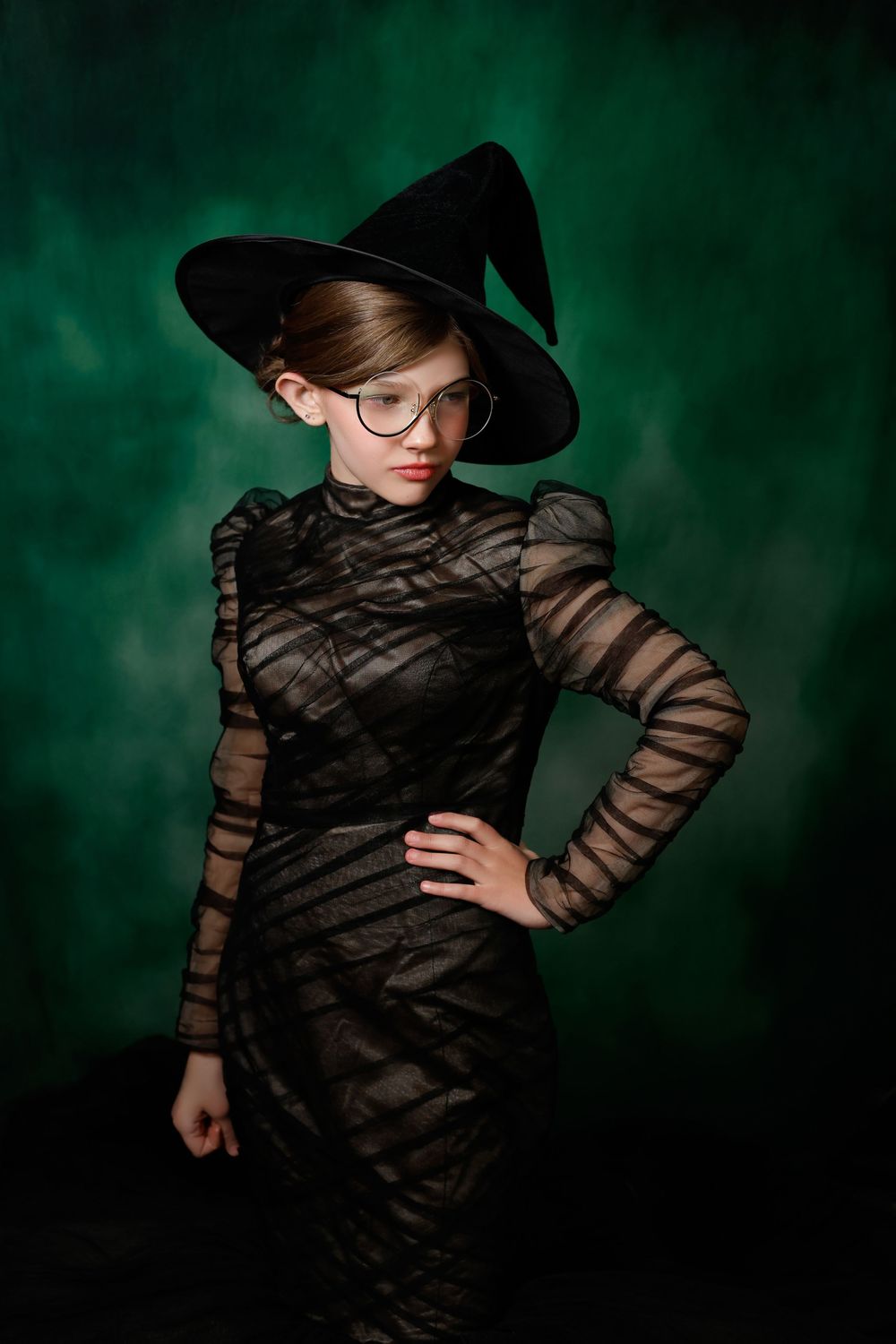 Young girl in black witch costume with pointed hat and glasses poses against a dark green background.