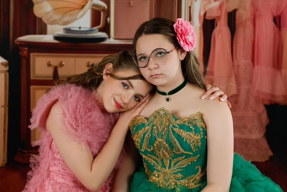 Two young women pose together, one in a pink feather jacket and one in an emerald green embroidered gown with a floral headpiece.