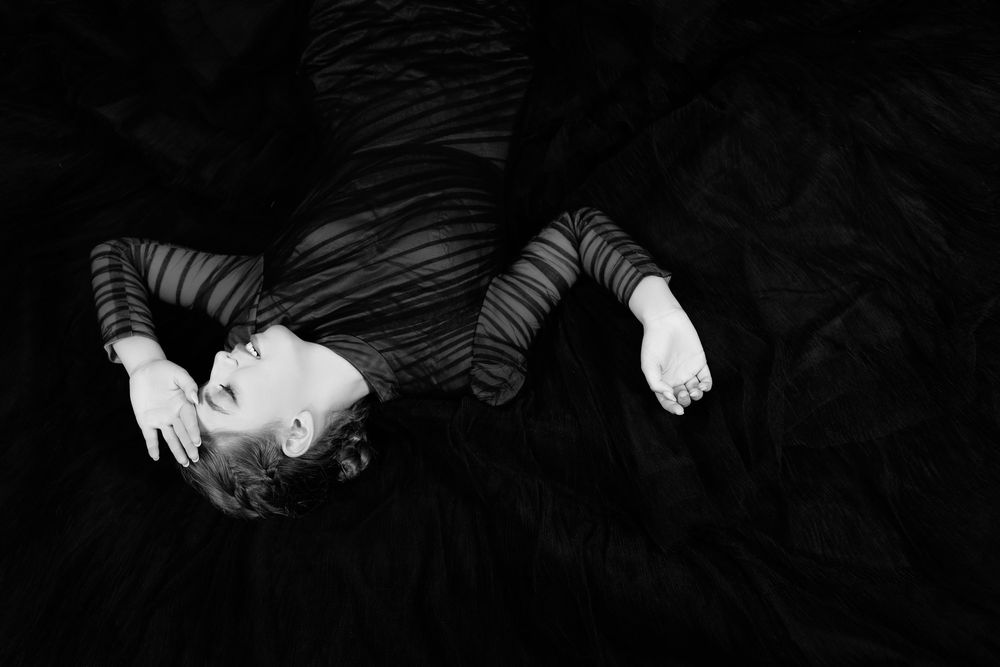 Black and white artistic photo of a person lying back dramatically on dark flowing fabric.