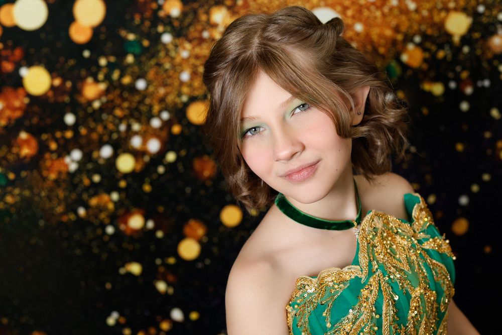 Close-up portrait of a girl in a green and gold gown against a sparkling bokeh background.