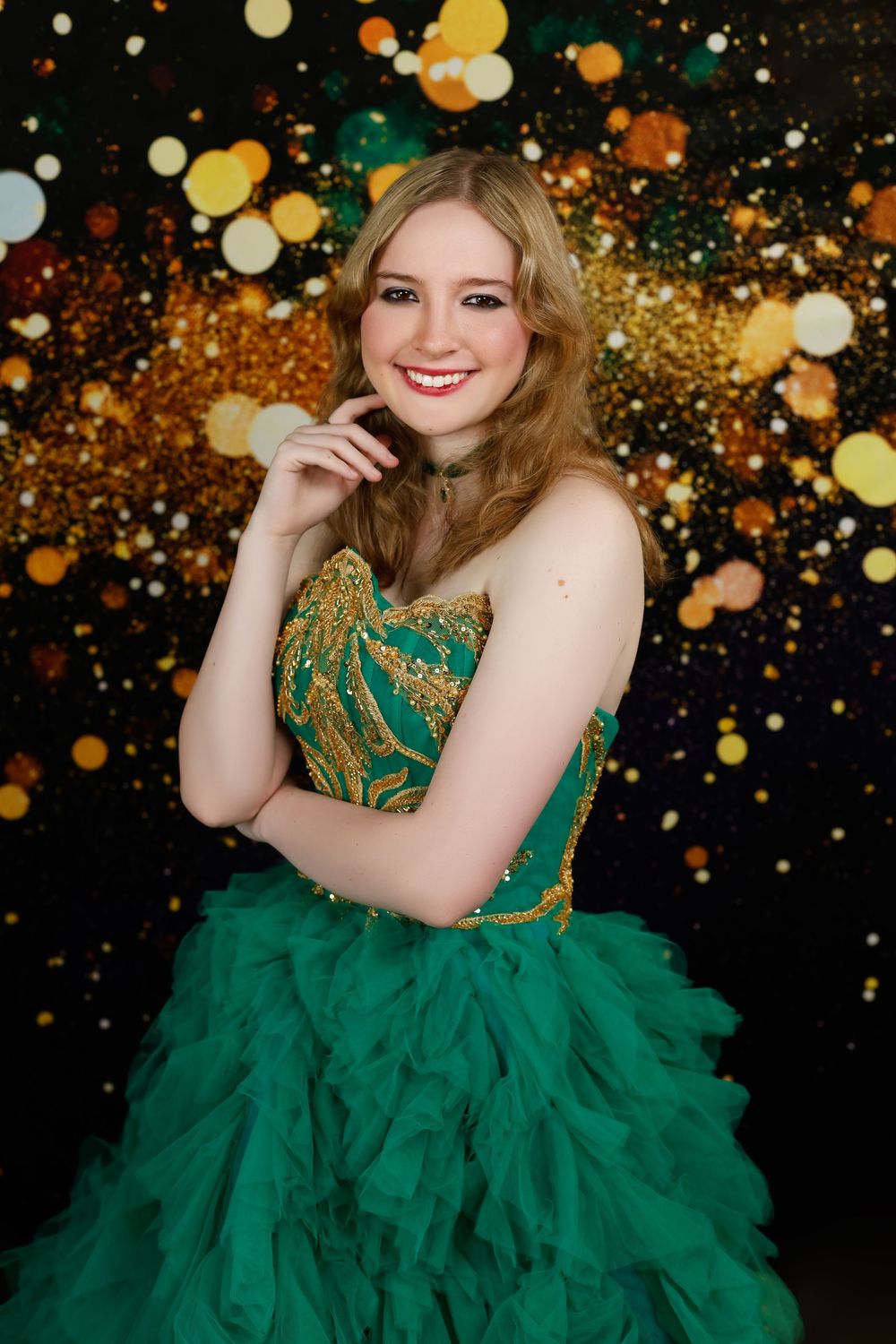 Smiling young woman in green and gold strapless ball gown poses against a golden bokeh backdrop.