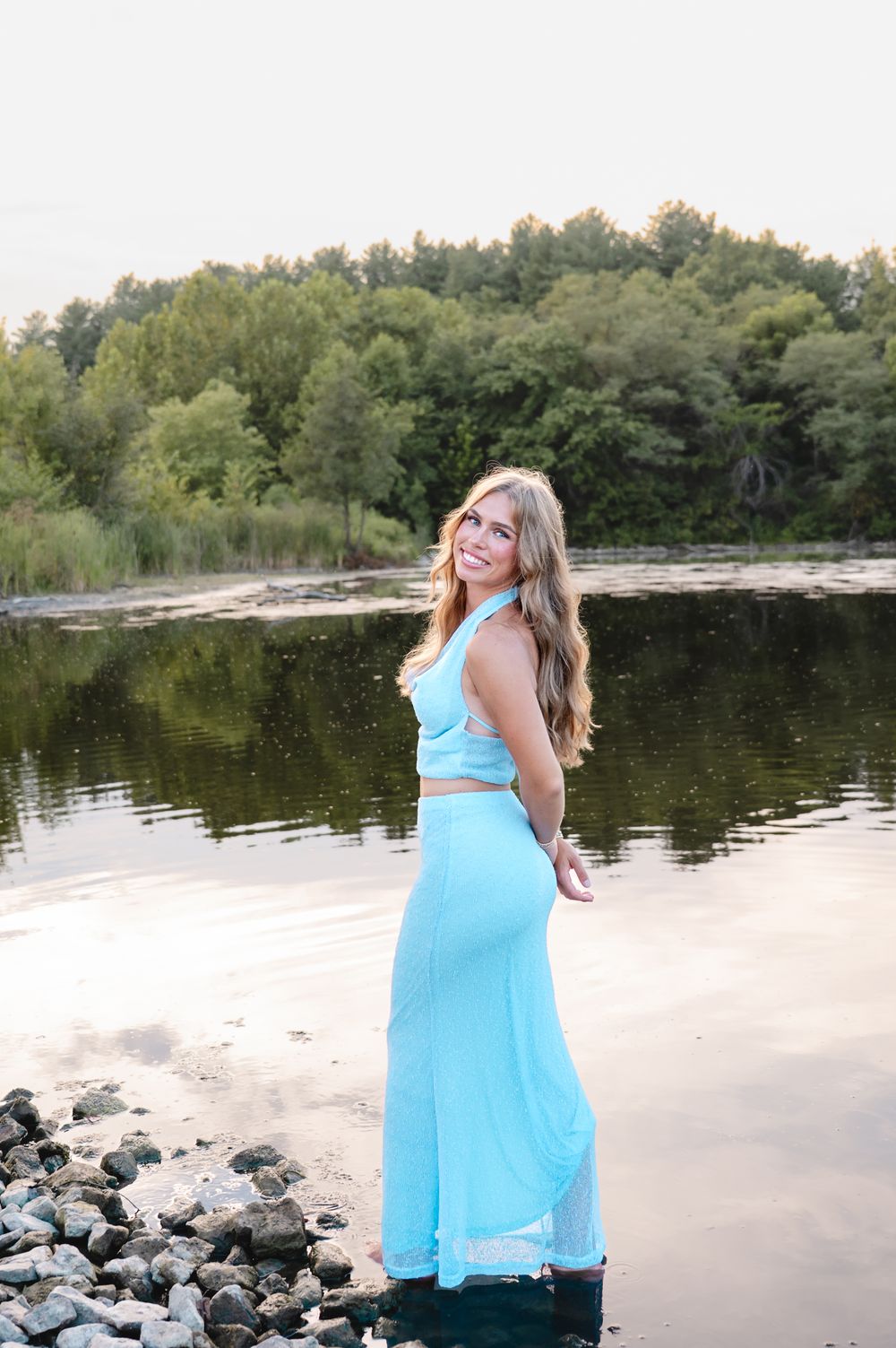 Woman in blue dress poses gracefully along a serene lakeshore with trees reflecting in the water.