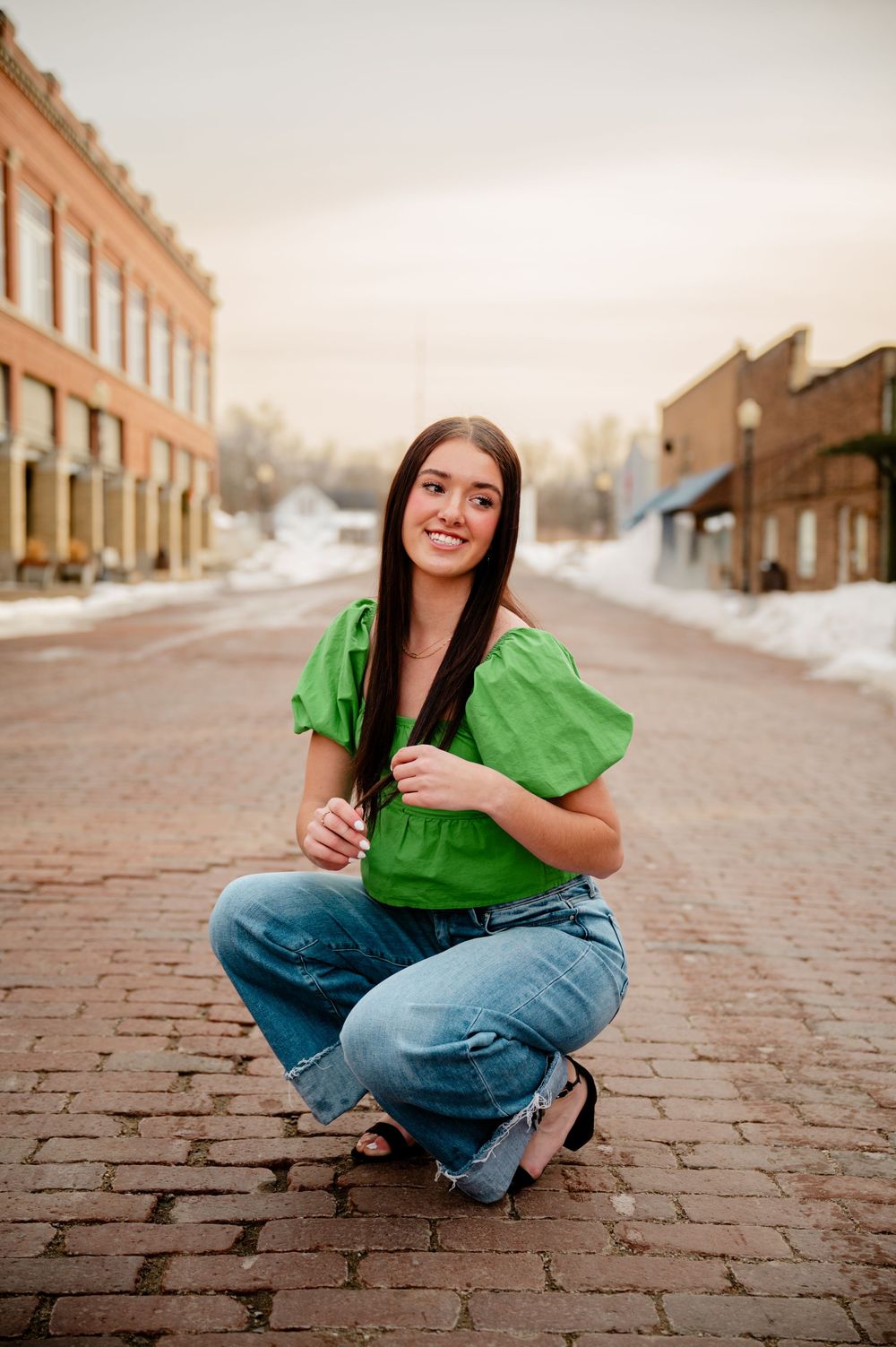 Young woman in green top and jeans crouching on brick street with snowy buildings backdrop at sunset.
