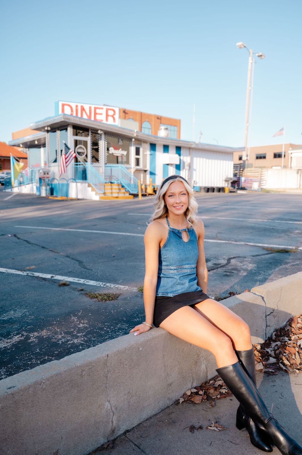 Graduate in black boots poses on concrete barrier with vintage diner in background.