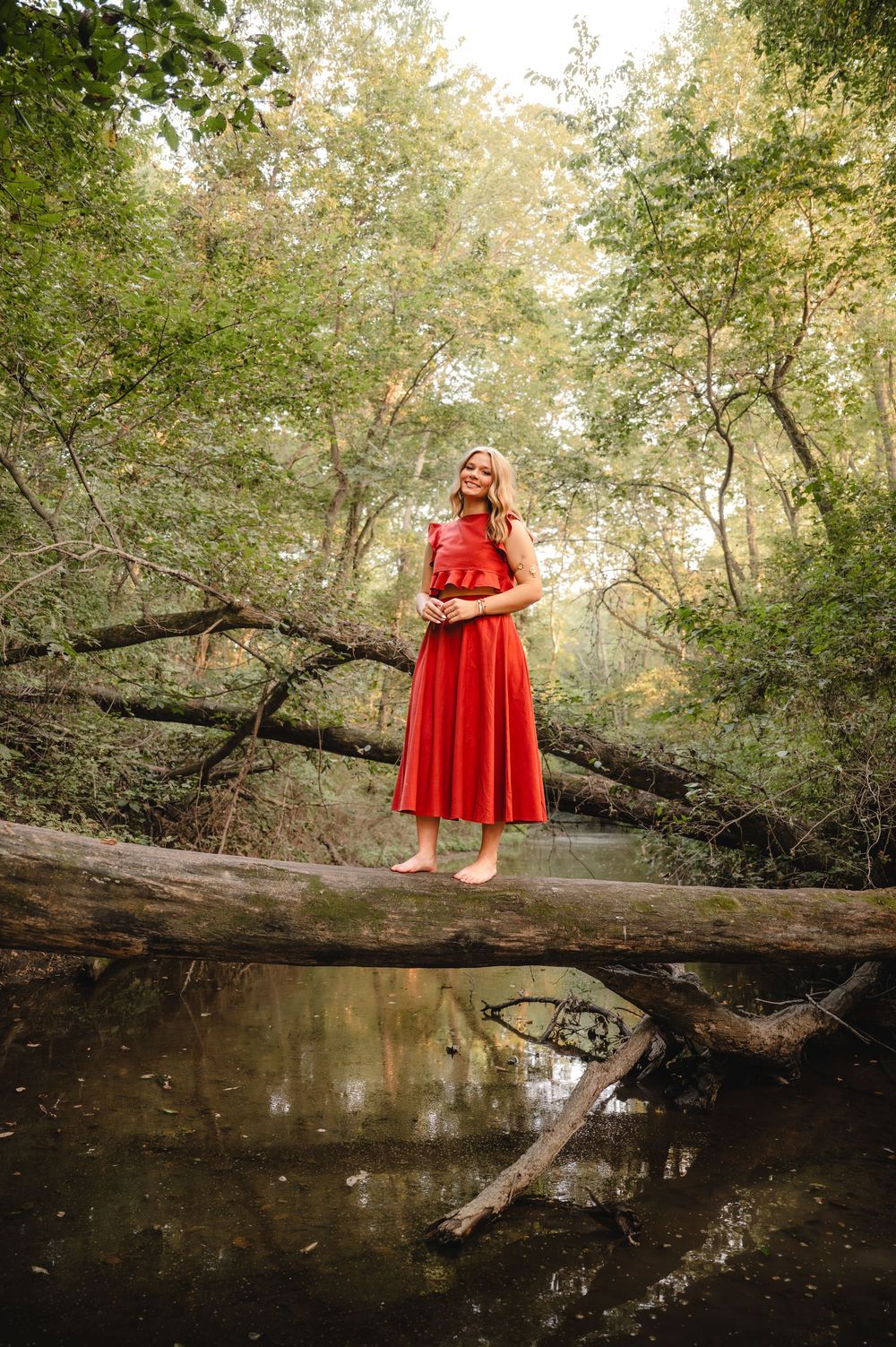 A dreamy portrait of someone in a flowing red dress standing on a fallen log over a creek.
