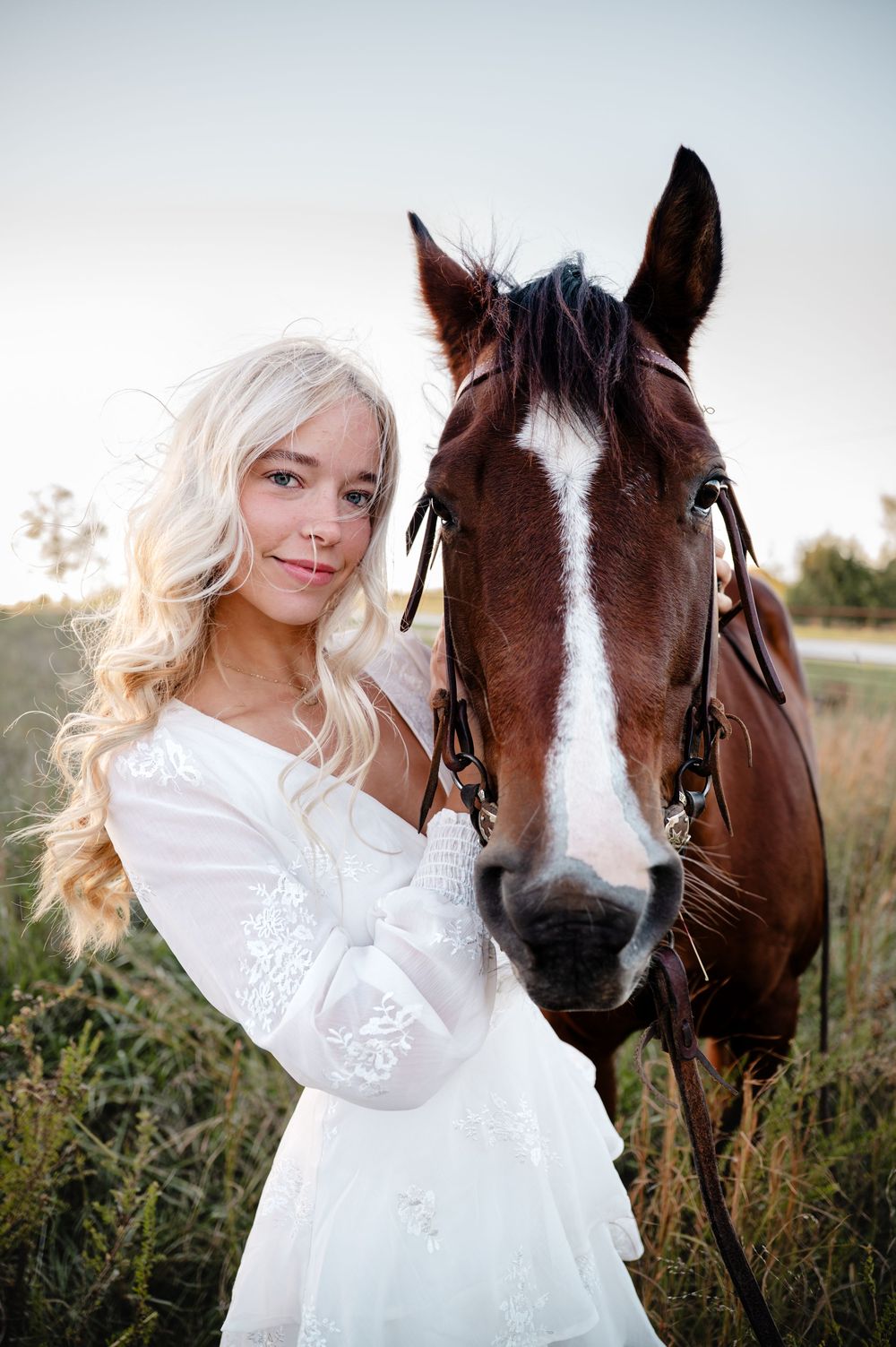 Person in white dress poses with brown and white horse in grassy field.
