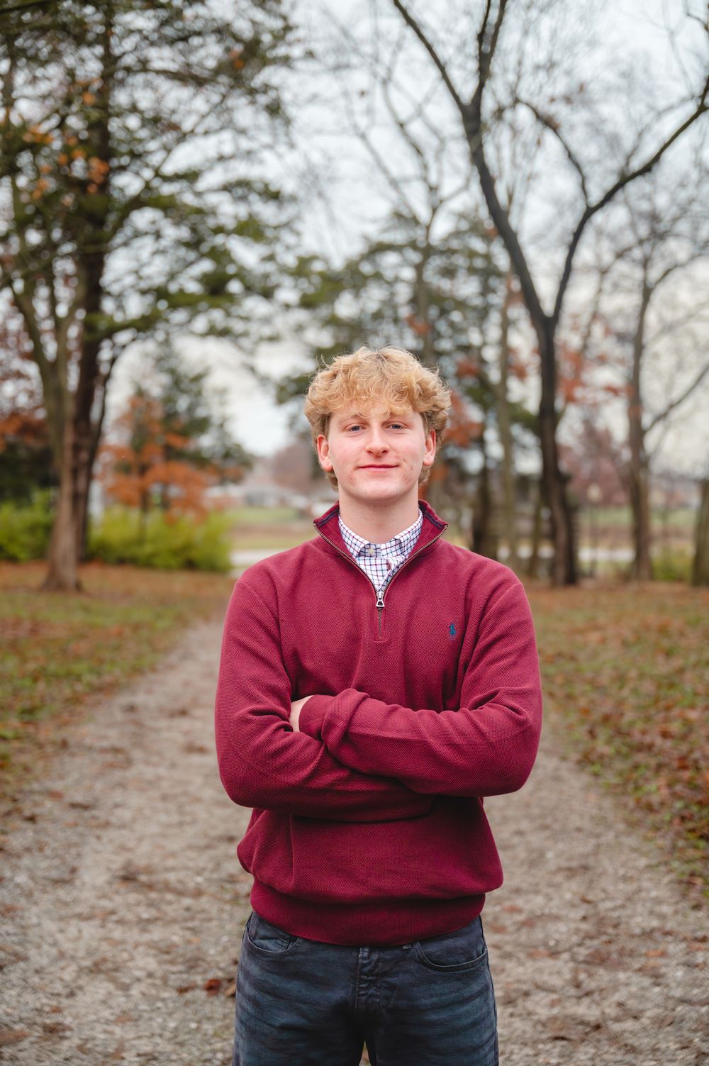 Student in burgundy sweater poses on autumn path with fallen leaves.