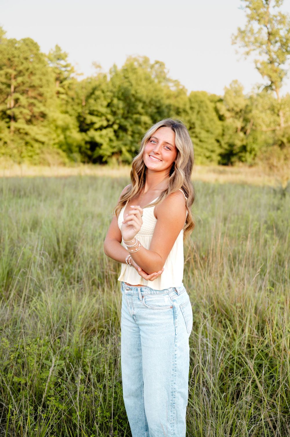 Woman in white top and jeans poses playfully in a sunny field with tall grass and trees in background.