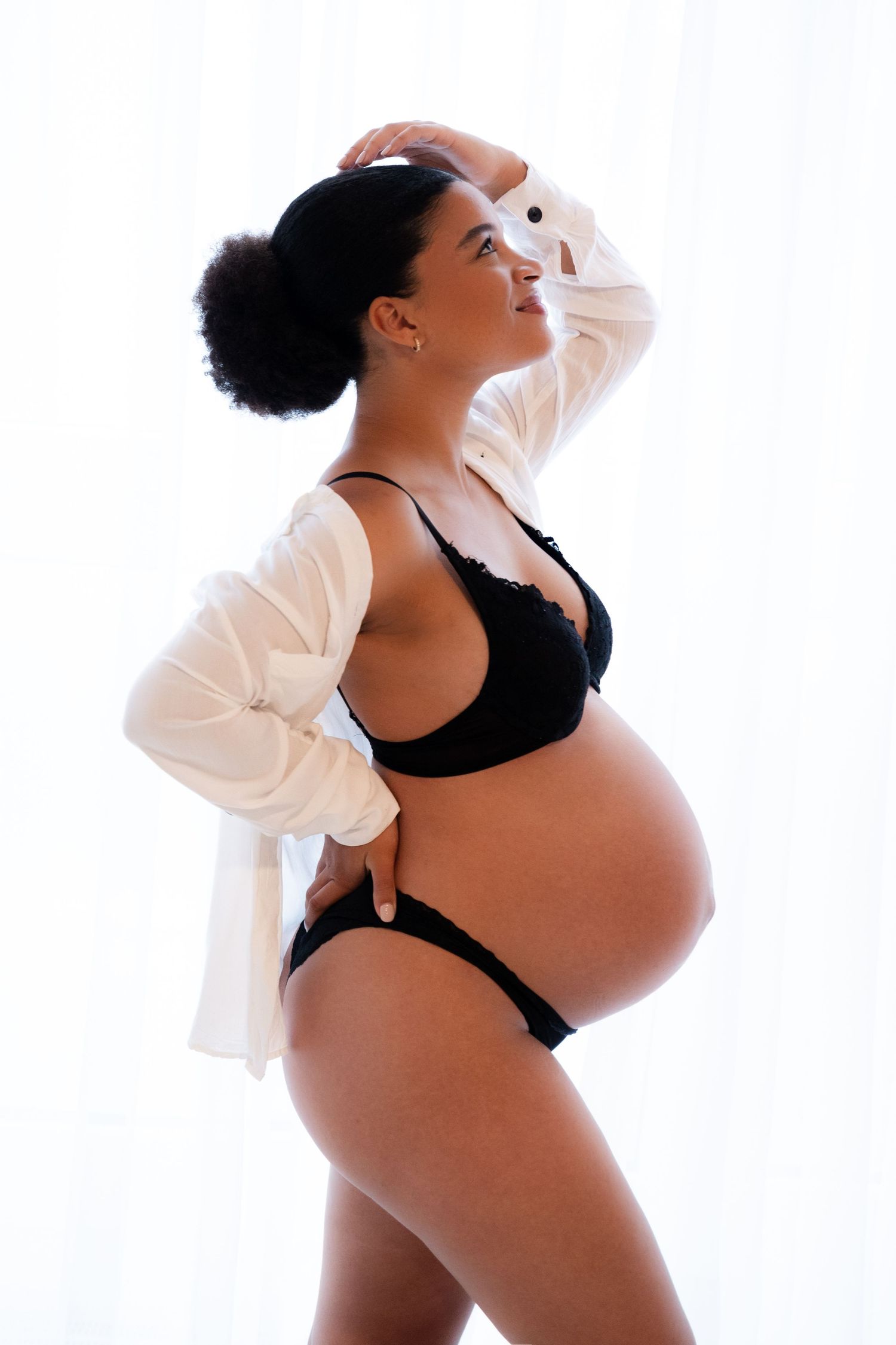 Pregnant woman in black lingerie and white robe posing confidently, hand raised to head, against white curtains.