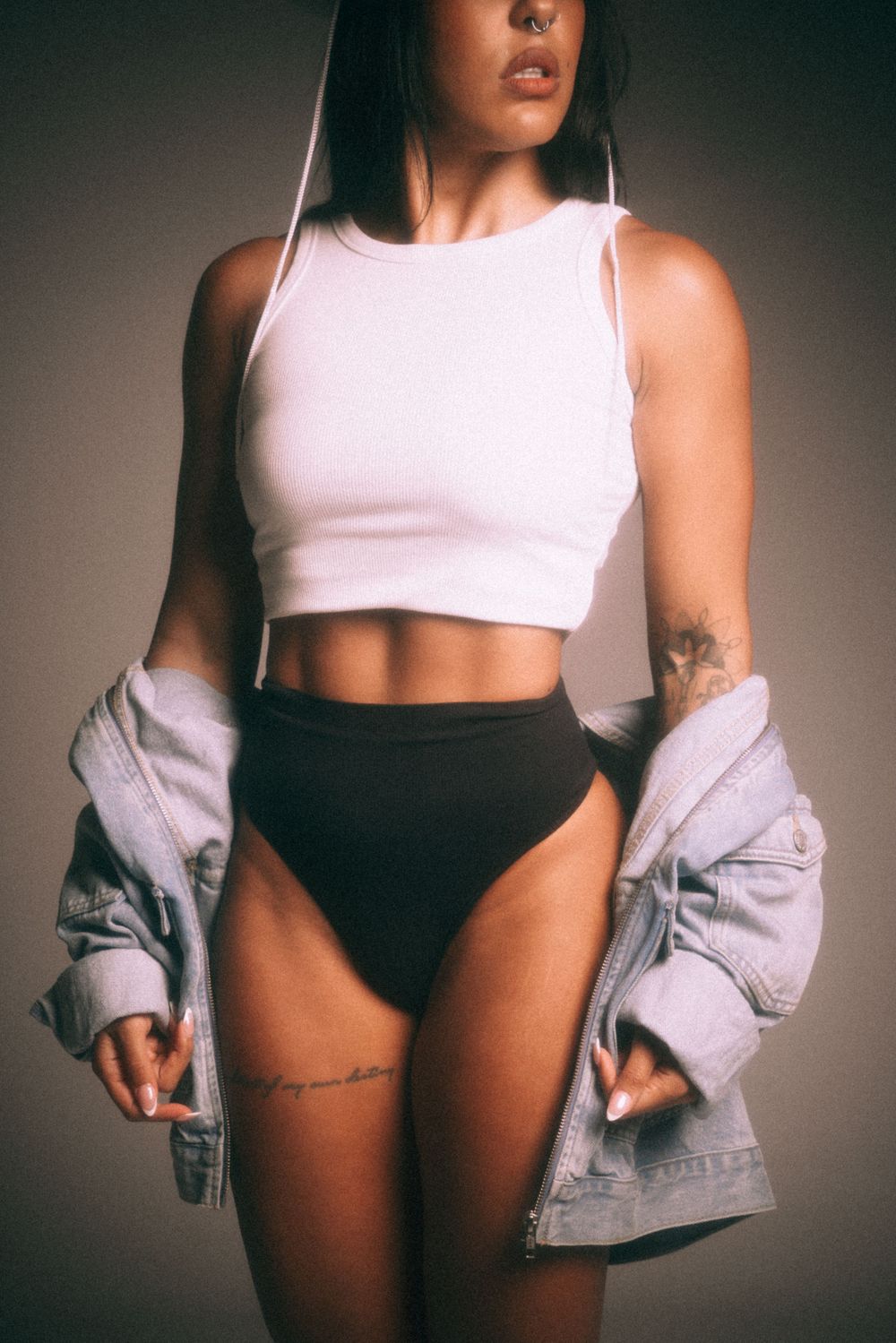 Fashion model poses in white crop top and black bottoms with denim jacket in moody studio lighting.