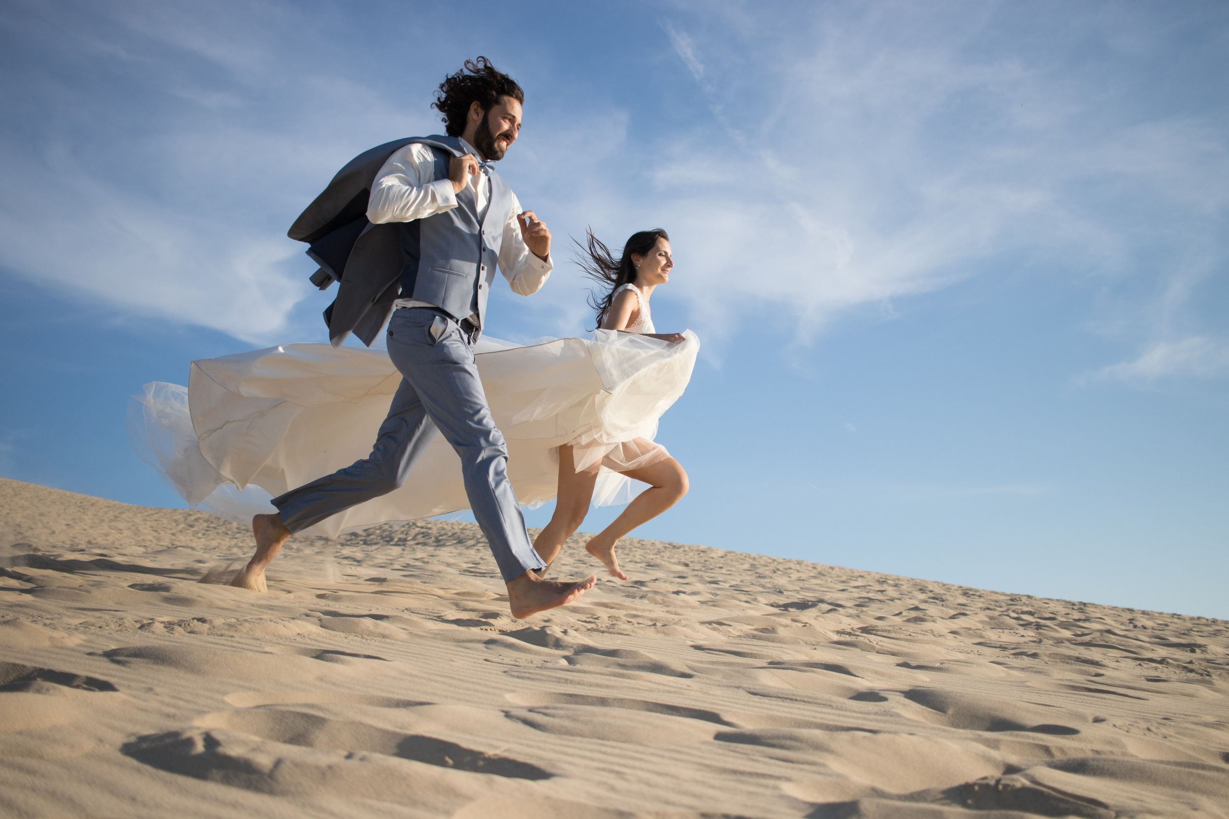 Two people running joyfully across sand dunes in formal attire with ocean in background.
wedding photo sevilla
mariage à la plage