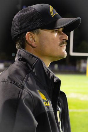Football coach wearing a black cap and jacket stands on the sideline during a night game.