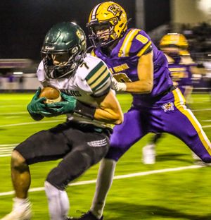 Green-uniformed football player catches the ball while being tackled by a purple-jersyed defender at night.