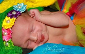 A sleeping newborn baby wearing a colorful floral headband on vibrant rainbow fabric.