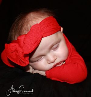 A peaceful sleeping baby in red outfit with a red bow headband on dark background.