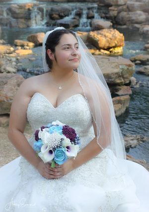 Beautiful bride in a strapless lace wedding gown holding a blue and purple bouquet near a waterfall.