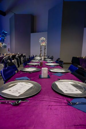 Elegant banquet table set with silver plates and blue napkins on a purple tablecloth with glowing centerpieces.
