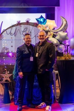 Two men in formal attire posing together in front of a colorful balloon arch at a gala event.