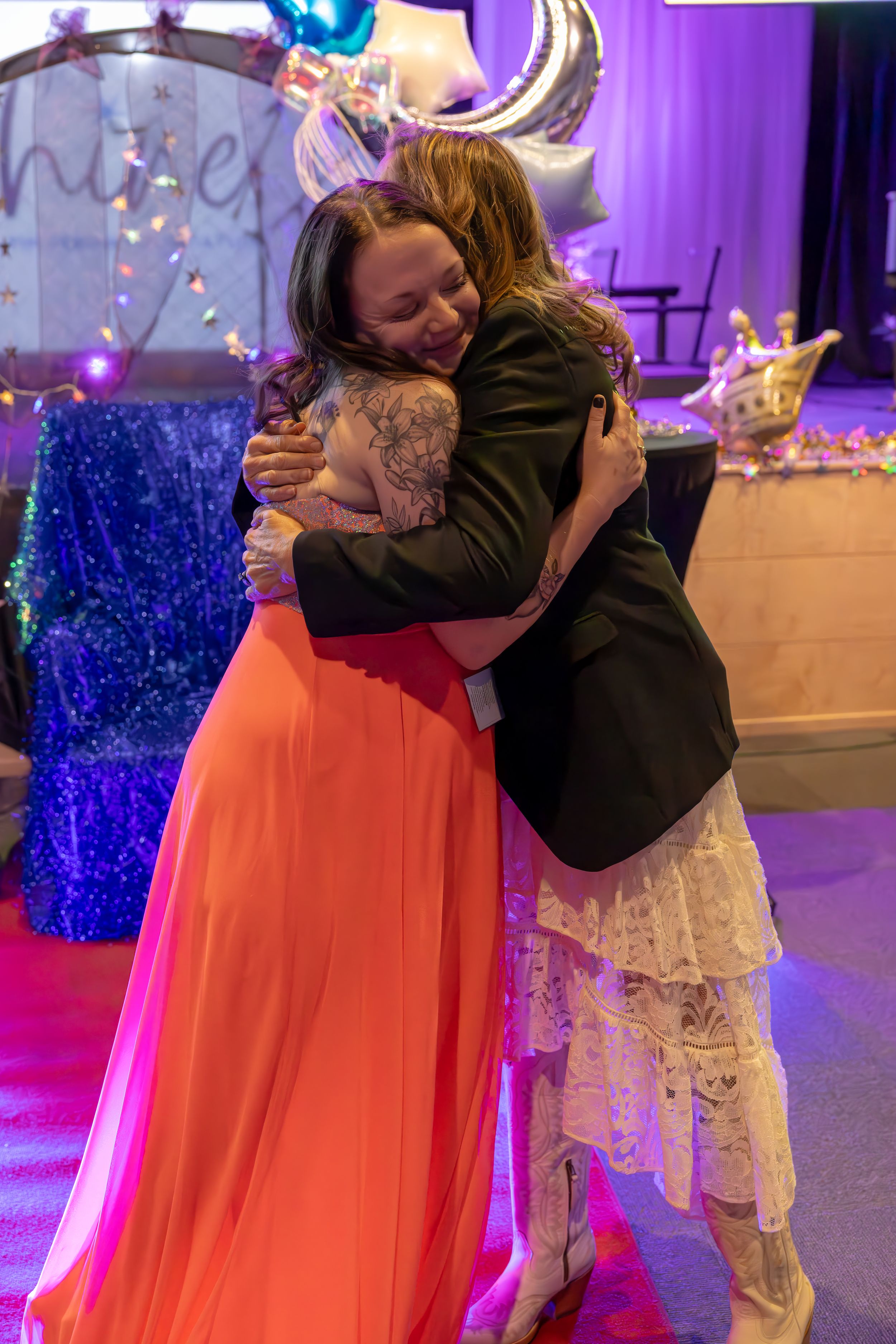 Two women share warm embrace at Night to Shine prom event with festive balloon decorations.