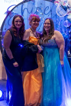 Three women in formal gowns smiling together in front of a decorative circular photo backdrop.