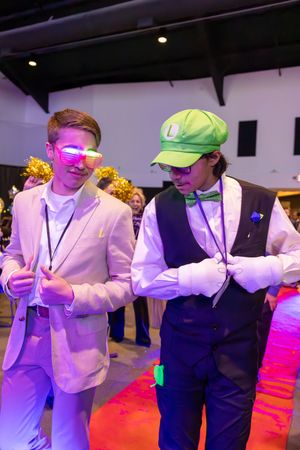 Two young men in formal attire dance at prom, one wearing LED glasses and green Luigi cap.