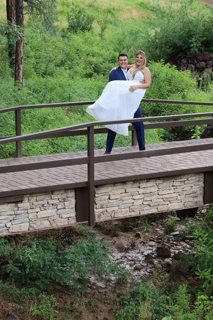 Groom carrying bride in wedding dress on a wooden bridge surrounded by lush green forest.
