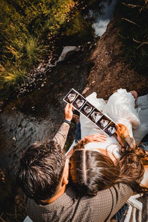 Couple sitting outdoors by a stream viewing ultrasound photos, aerial view of pregnancy announcement.