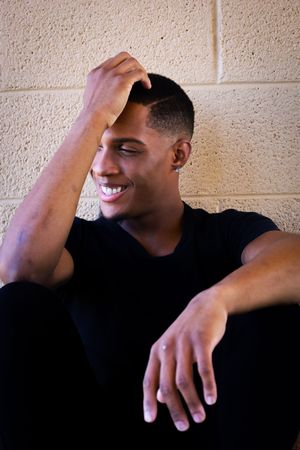 Smiling young man in black shirt leaning against a tan brick wall with hand on his head.