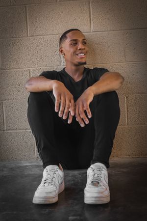 Confident young man in black outfit and white sneakers sitting against a brick wall smiling.