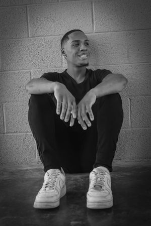 Black and white portrait of a smiling young man sitting against a brick wall in white sneakers.