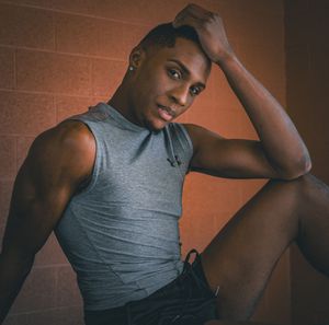 Athletic young man in gray sleeveless top posing against an orange brick wall with hand in hair.