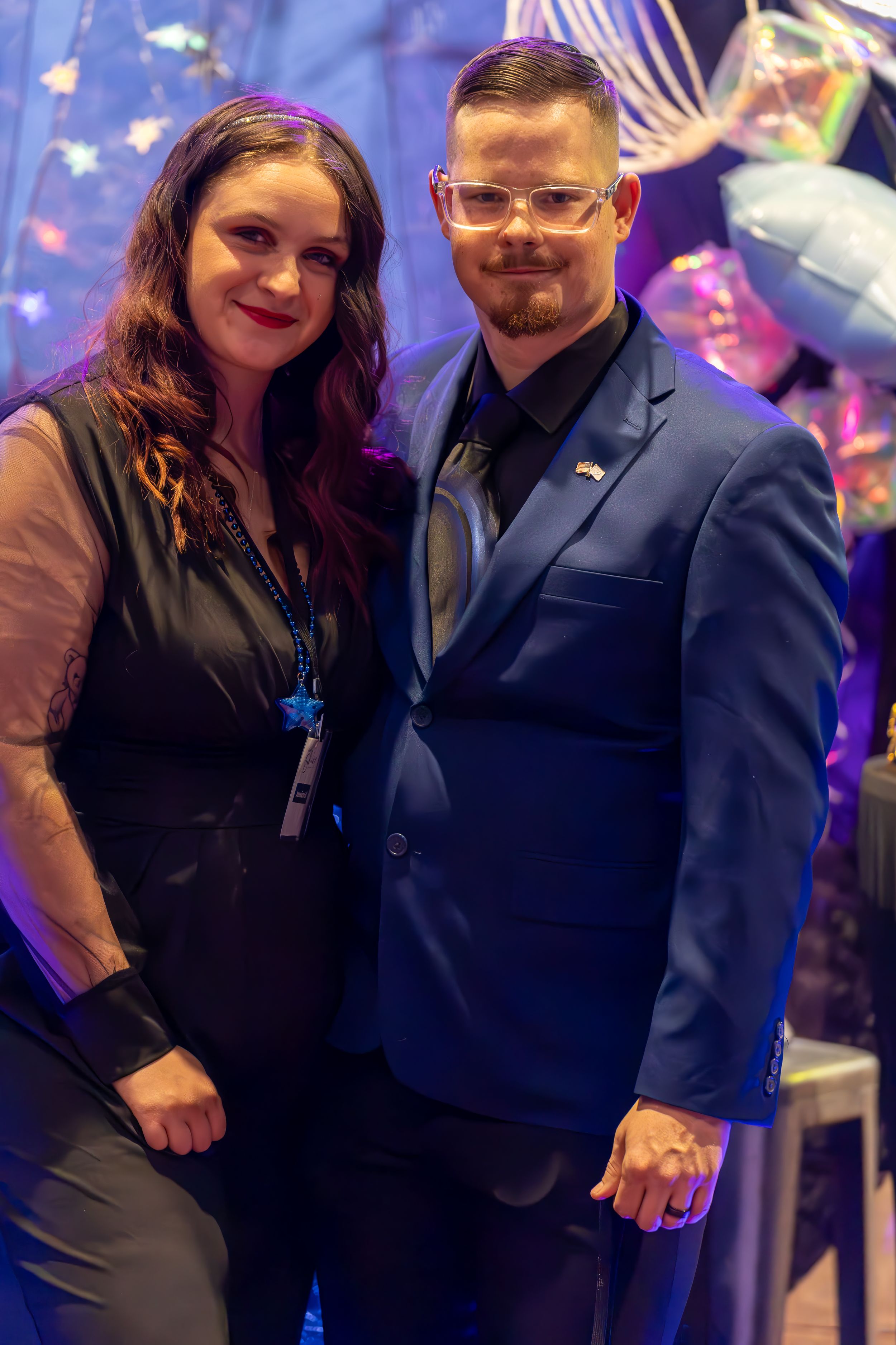 Elegant couple posing together at a formal event with colorful balloon decorations behind them.