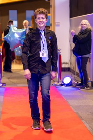 Smiling teenage boy walking confidently down a red carpet at a formal event.