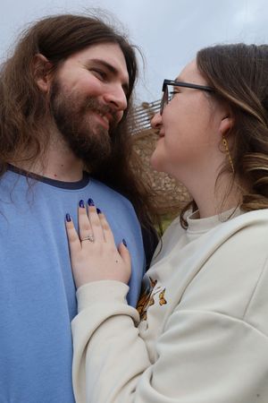 Close-up of smiling couple nearly kissing outdoors, woman showing engagement ring with blue nails.