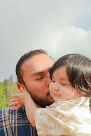 Bearded father lovingly kissing young toddler daughter on cheek against cloudy sky.