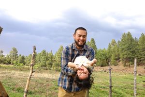 Laughing father playfully holding young child upside down in rural field with pine trees.
