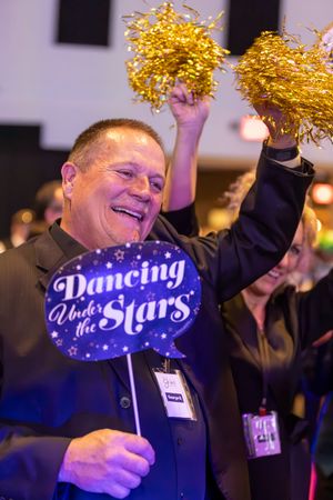 Cheerful man holds Dancing Under the Stars sign and gold pom-poms at festive gala event.