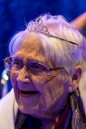 Elderly woman wearing sparkling tiara and glasses smiles joyfully at Night to Shine prom event.