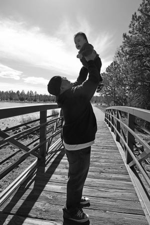 A father lifts his laughing baby high on a wooden bridge boardwalk in black and white.
