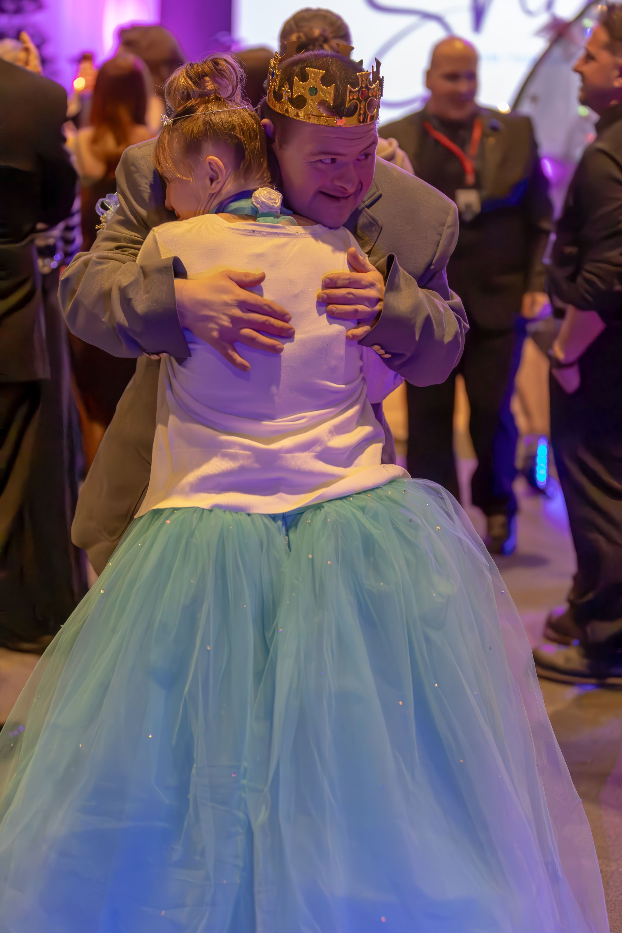 Two people sharing a warm hug on a dance floor, one wearing a crown and tutu skirt.