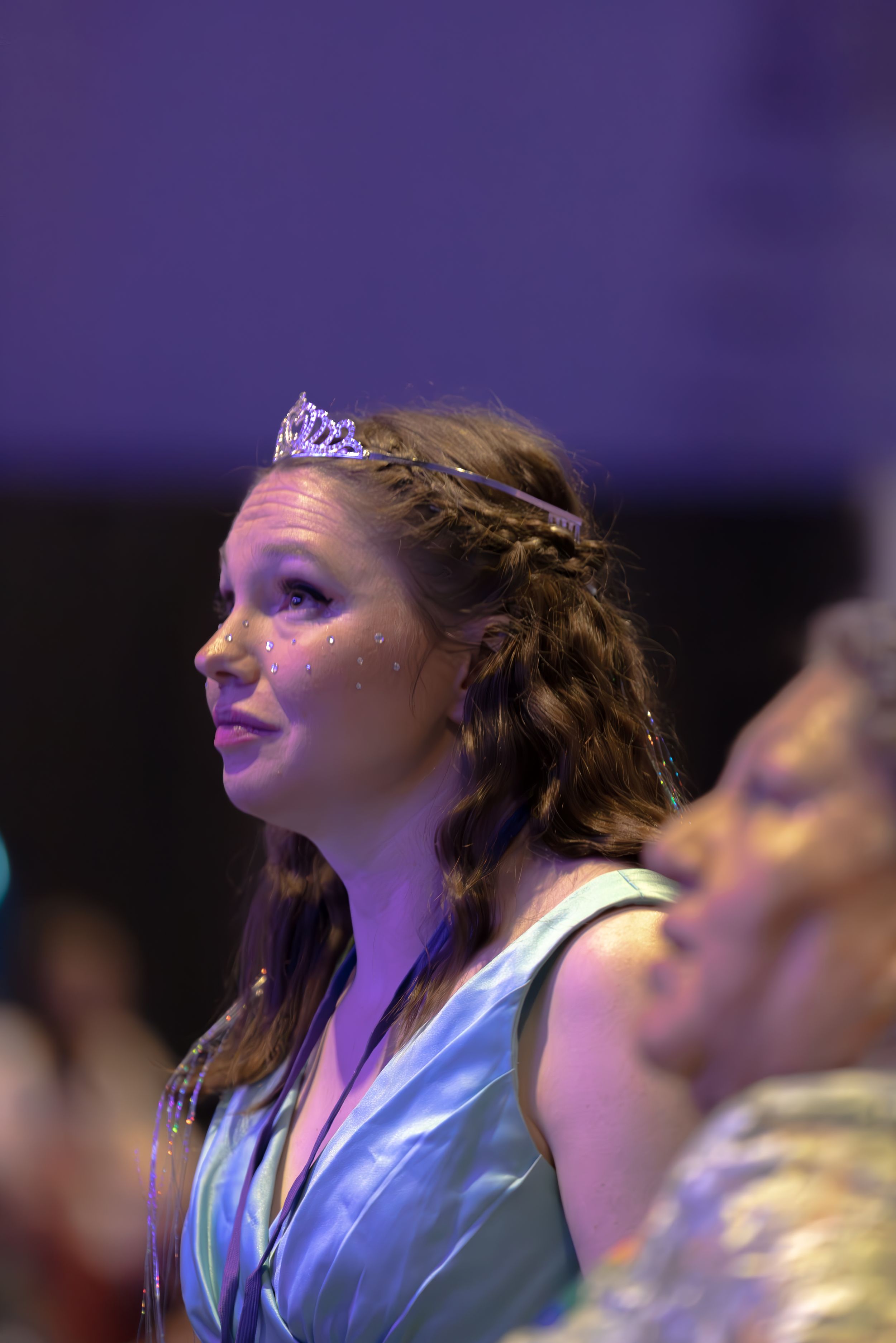 A young woman wearing a tiara and glitter at a formal event under purple lighting.
