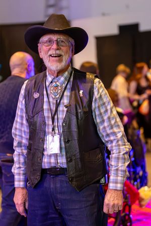 Smiling senior man in cowboy hat, vest, and bolo tie poses at festive indoor gala event.