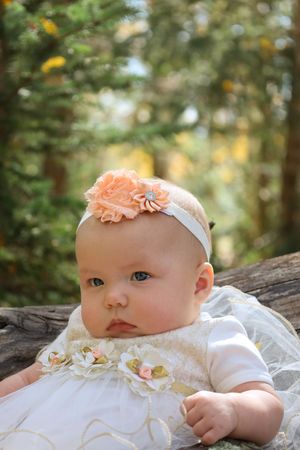 An alert baby in a white floral dress with peach headband rests on a log outdoors.