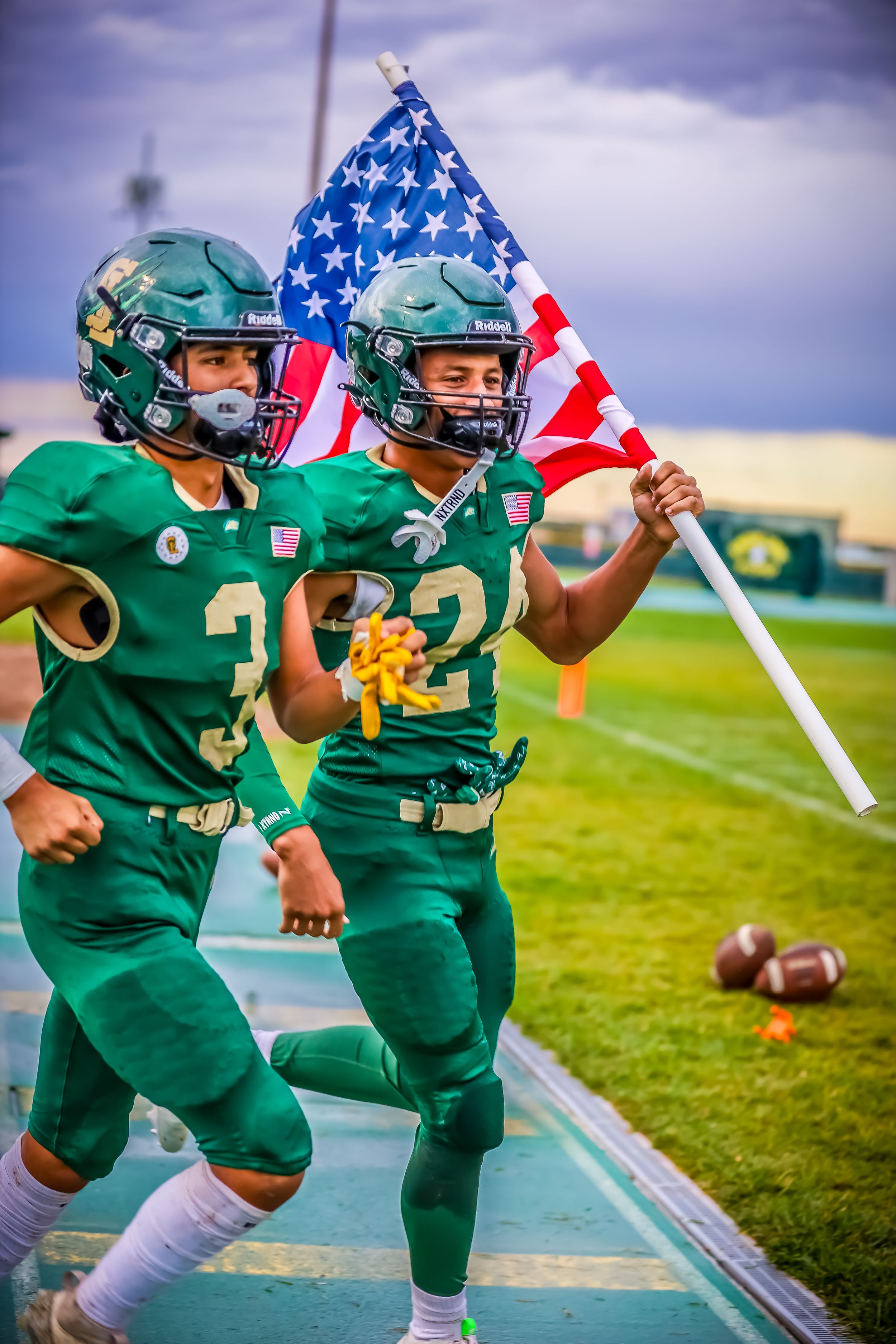 Two football players in green uniforms running onto field carrying an American flag.