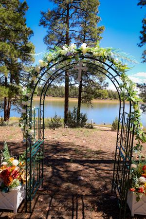 Floral decorated metal wedding arch with cross near a lake surrounded by pine trees.