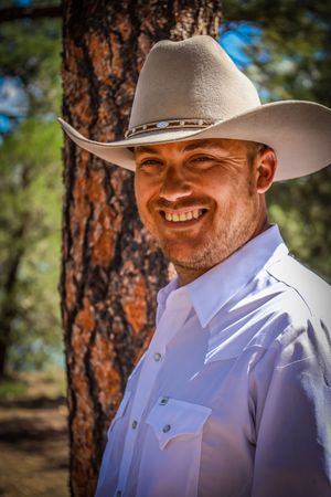 Smiling man in white shirt and cowboy hat poses outdoors near pine tree.