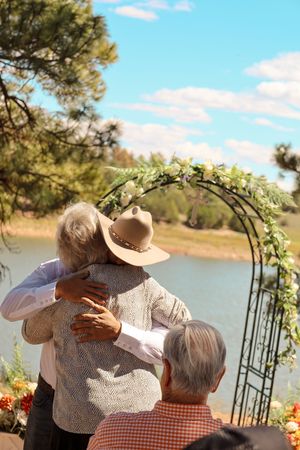 Couple embraces under floral arch by lake at outdoor wedding ceremony.