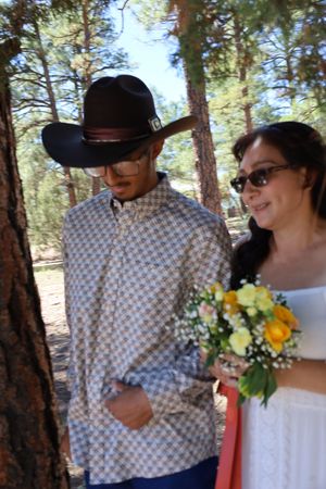 Man in cowboy hat and woman holding yellow bouquet stand by pine tree.