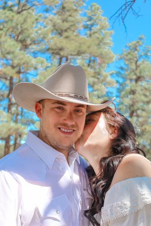Groom in cowboy hat smiles as bride kisses his cheek outdoors at wedding.